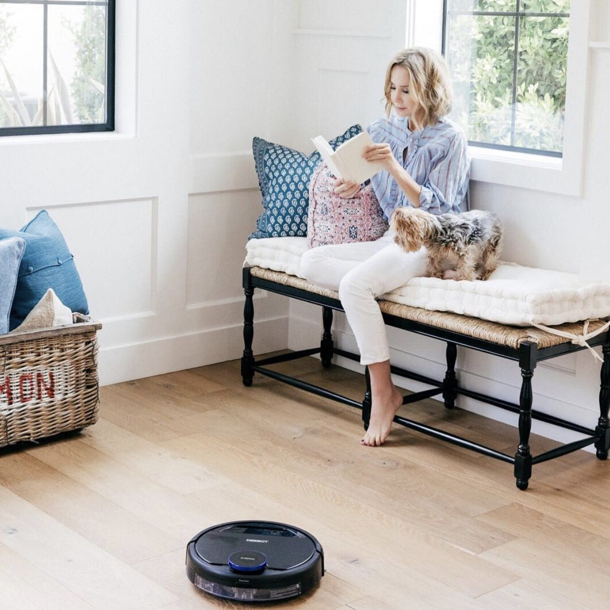 Woman sitting on bench reading a book with a small dog next to her, while a robotic vacuum cleans the floor