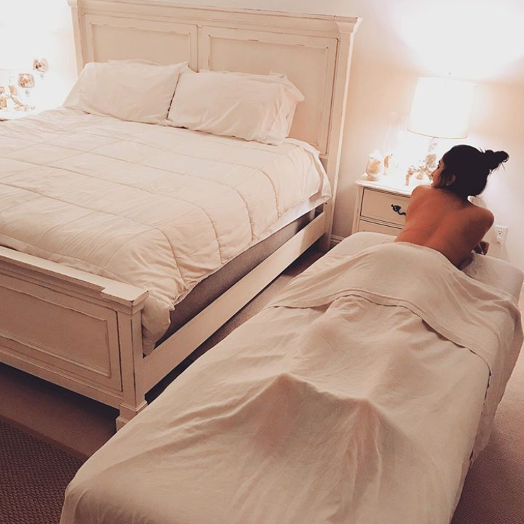 Woman lying on massage table and smiling, with bed and other bedroom furniture around her